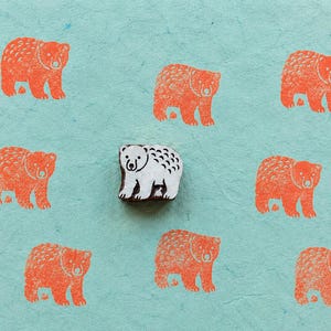 May include: A small, rectangular wooden block stamp featuring a carved bear design, positioned in the centre of a light blue surface. Surrounding the stamp are seven orange ink impressions of the same bear, creating a repeating pattern.