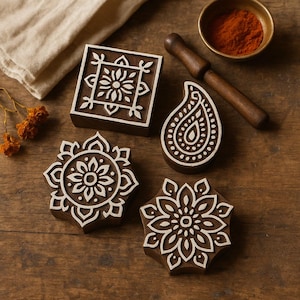 May include: A collection of intricately carved wooden printing blocks in various floral and geometric designs. The blocks are dark brown with white detailing, alongside a small bowl of red powder and a wooden pestle. The arrangement is on a wooden surface.