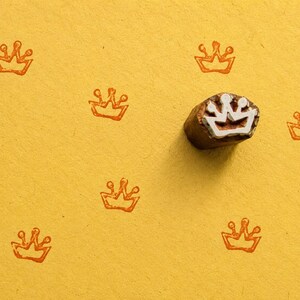 May include: A small, brown, carved wooden stamp with a white crown-shaped relief pattern on its face, positioned next to several orange ink-stamped crown impressions scattered across a textured, solid yellow paper surface.