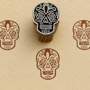 May include: A wooden block stamp featuring a carved sugar skull design with intricate floral patterns. The stamp is positioned above three brown ink impressions of the same skull design, displaying detailed eye sockets and teeth.