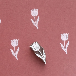 May include: A wooden stamp featuring a white tulip design, alongside several stamped white tulip images, set against a dusty rose backdrop. This is a craft supply for creating floral patterns.