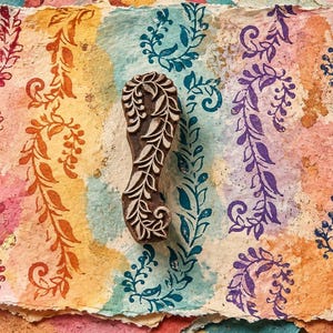 May include: A dark brown wooden block print stamp featuring an intricate carved leaf and vine pattern. The stamp is positioned vertically on a textured, multi-coloured paper background that displays several stamped impressions of the same design.