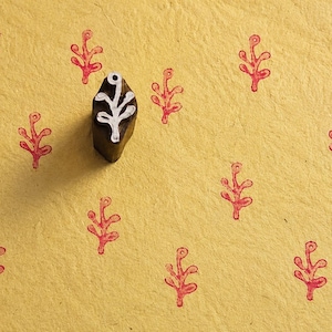 May include: A dark brown wooden block stamp with a white carved plant motif on its base, positioned on a textured yellow surface that features multiple red stamped impressions of the same plant design in a repeating pattern across the paper.