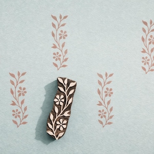 Puede incluir: Un bloque de madera con un diseño floral, con flores y hojas blancas sobre un fondo marrón oscuro. El bloque está rodeado por un patrón repetitivo de ramitas florales en un color rosa apagado sobre un fondo azul claro. Ideal para proyectos de artesanía.