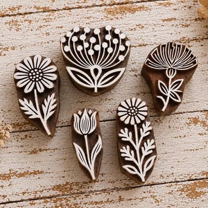 May include: Five dark brown wooden printing blocks featuring carved white floral designs. The set includes various flower shapes such as daisies, a tulip, and a large rounded bloom, each with detailed stems and leaves for textile or paper stamping.
