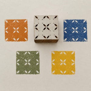 May include: A square wooden printing block with a carved four-petal floral pattern, positioned above four square ink prints in orange, blue, olive green, and yellow, each featuring the same repeating four-petal floral design and small dots.
