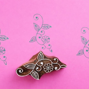 May include: A wooden block with a floral design and three teal floral patterns on a pink background. The block is brown with white floral details. The teal patterns include leaves, swirls, and flowers.