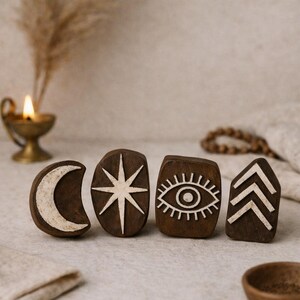 May include: Four dark brown wooden blocks with white painted designs: a crescent moon, an eight-pointed star, an eye, and three upward-pointing chevrons. The blocks are arranged on a neutral-toned surface.