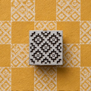 May include: A square wooden printing block featuring a carved geometric pattern of repeating diamonds and crosses. The block is positioned over a yellow and white checkered surface that displays the same intricate, stamped geometric design.