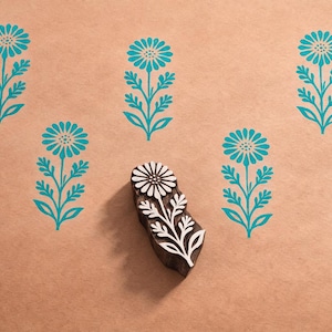 May include: A wooden block with a floral design, featuring a daisy-like flower and leaves, is surrounded by teal floral prints on a tan background. The block is used for printing and creating patterns.