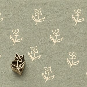 May include: A wooden block stamp featuring a carved four-petal flower design with leaves, positioned on a sage green textured surface that displays a repeating pattern of the same white flower motif printed across the background.