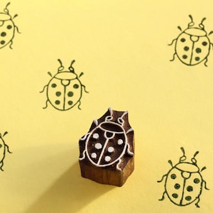 Könnte beinhalten: Ein Holzstempel mit einem Marienkäfer-Design, mit weißem Umriss und Punktdetails, liegt auf einem gelben Hintergrund mit mehreren Marienkäfer-Illustrationen. Der Stempel ist dunkelbraun, und die Marienkäfer-Illustrationen sind schwarz.