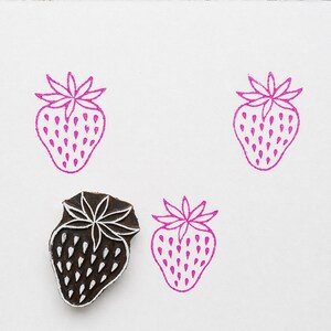 以下が含まれることがあります： 白と茶色のデザインの木製イチゴスタンプと、白い紙に描かれた3つのピンク色のイチゴの輪郭。スタンプは、星型のトップと小さなドットが付いたイチゴの詳細なデザインです。スタンプされたイチゴは、シンプルな線画スタイルです。