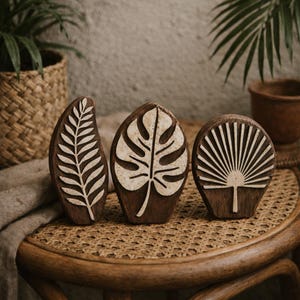 May include: Three wooden leaf-shaped stamps with white designs. The stamps are in the shape of a fern, a monstera leaf, and a fan palm. They are displayed on a woven chair with a neutral background.