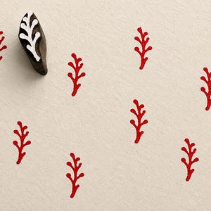 May include: A dark brown wooden block stamp with a carved white branch design, positioned next to eight red stamped branch impressions. The stamp features a textured surface and a distinct organic shape, creating a repeating pattern of red branches.