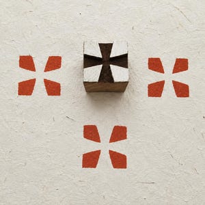 Pode incluir: Um carimbo de bloco de madeira quadrado com um design de cruz esculpido com tinta branca na superfície. O carimbo está posicionado acima de três impressões idênticas em forma de cruz laranja numa superfície de papel texturizado esbranquiçado.