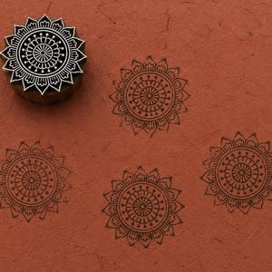 May include: A circular wooden printing block featuring a detailed floral mandala pattern, shown alongside four dark brown ink impressions of the same design stamped onto a textured, terracotta-colored surface. The block has a carved relief.