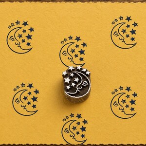 May include: A wooden printing block featuring a carved crescent moon with a face and surrounding stars, positioned on a mustard yellow paper surface stamped with six identical dark blue ink impressions of the same moon and star design.