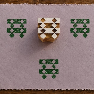 Könnte beinhalten: Ein quadratischer Holzdruckblock mit einem geometrischen Rautenmuster liegt auf einem hellvioletten, strukturierten Papier. Drei grüne Tintenabdrücke desselben geometrischen Designs sind auf das Papier gestempelt und dreieckig um den Block angeordnet.