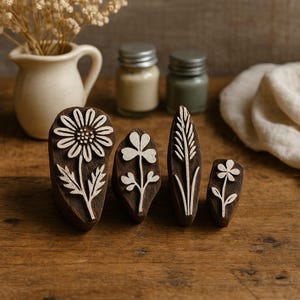 May include: Four wooden floral stamps with white designs. The stamps feature a daisy, a clover, a wheat stalk, and a small flower. The stamps are arranged on a wooden surface, with small jars and a vase of dried flowers in the background.