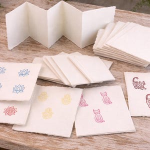 May include: Handmade paper products, including blank cards and a concertina booklet. The cards display stamped designs in blue, red, and yellow, such as lotus flowers, hands, and cat illustrations. The paper has a textured, natural appearance.