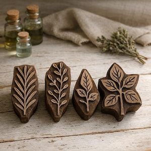 May include: Four wooden leaf-shaped stamps with intricate carved designs, arranged on a rustic wooden surface. The stamps are various shapes, including a fern, a sprig, and a leaf. Small glass bottles with cork stoppers are in the background.