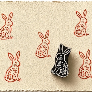 May include: A black rubber stamp shaped like a sitting rabbit with floral details, positioned on a textured cream-coloured paper. Four orange ink impressions of the same rabbit design are stamped around the physical stamp on the paper surface.