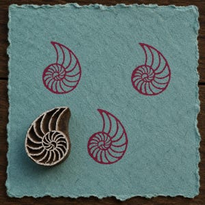 Op de afbeelding: Een vierkant stuk lichtblauw gestructureerd papier met gescheurde randen met drie magenta nautilusschelpafdrukken en één houten stempel met een bijpassend nautilusschelpontwerp geplaatst aan de linkerkant van het papier.