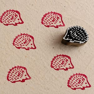 Puede incluir: Un pequeño sello de goma negro tallado con forma de erizo, colocado junto a seis impresiones en tinta roja del mismo diseño. El sello presenta un patrón detallado y estilizado de púas y un hocico puntiagudo, creando un motivo animal repetido.