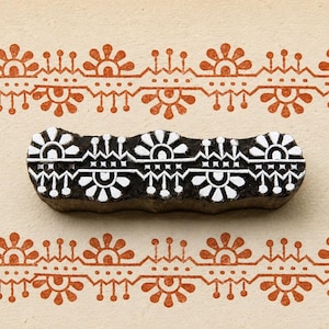 Könnte beinhalten: Ein Holzblock mit einem floralen Design in Weiß und Schwarz, der zum Drucken verwendet wird. Der Block befindet sich auf einem beigefarbenen Hintergrund, mit einem sich wiederholenden orangefarbenen Blumenrand oben und unten.