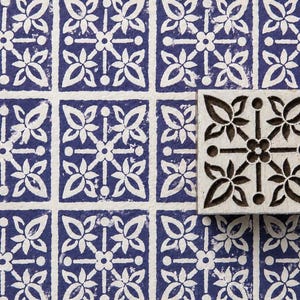 May include: A square wooden printing block featuring a carved floral geometric pattern, positioned on a surface printed with a repeating blue and white grid design that matches the motif on the block itself, creating a cohesive visual display.