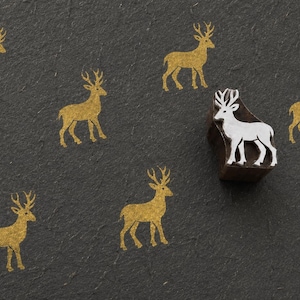 May include: A silver-colored metal stamp shaped like a deer, positioned on a dark gray textured surface. The surface features six stamped impressions of the same deer design in a shimmering gold color, arranged around the physical metal stamp.