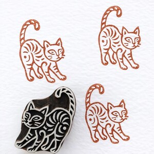 May include: A wooden block stamp with a cat design, alongside three stamped impressions. The stamp is black with a white cat design. The stamped images are in brown ink, showing a stylized cat with striped markings and a curled tail. The cat is depicted in a walking pose.