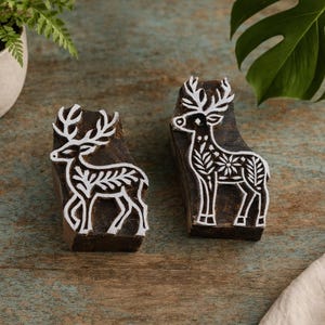 May include: Two wooden block stamps with white deer designs. The deer have detailed antlers and floral patterns on their bodies. The blocks are dark brown and sit on a rustic, teal-colored wooden surface. A small potted plant and a large green leaf are in the background.