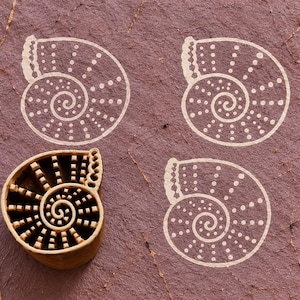May include: A carved wooden block in the shape of a spiral seashell with a series of raised rectangular ridges and a scalloped edge. Three white ink impressions of the same seashell design are stamped on the textured mauve surface nearby.