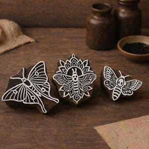May include: Three wooden butterfly stamps with white detailed designs. The stamps are arranged on a wooden surface, with a neutral background. Other items include small brown jars, a bowl, and a roll of twine.