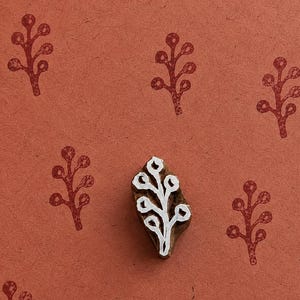 May include: A wooden block printing stamp featuring a carved branch motif with rounded leaves. The stamp is positioned on a textured terracotta-colored surface that displays several repeated red ink impressions of the same branch design.