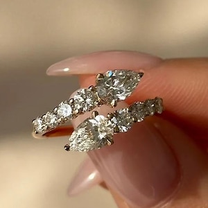 Puede incluir: Dos anillos de oro con piedras preciosas claras y brillantes. Un anillo presenta una piedra en forma de pera, el otro una serie de piedras más pequeñas. Los anillos son sostenidos por una mano con uñas cuidadas.