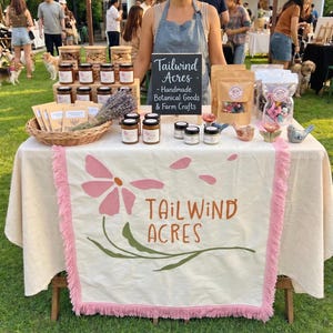 Könnte beinhalten: Ein Marktstand im Freien präsentiert handgemachte botanische Waren und Produkte vom Bauernhof. Gläser mit Konserven, verpackte Waren und Dekorationsartikel sind auf einem Tisch mit floralem Design und dem Text "Tailwind Acres" angeordnet.