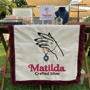 May include: A display of Matilda Crafted Silver jewelry. A cream-colored table runner with a burgundy fringe features an embroidered hand holding a necklace. The Matilda logo is displayed on a sign and the table runner.
