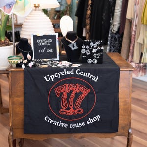 Peut inclure: Un chemin de table noir avec le texte blanc Upcycled Central creative reuse shop et un graphique rouge 777. Sur la table se trouvent des présentoirs à bijoux avec colliers et boucles d'oreilles, une lettre avec UPCYCLED JEWELS X 1 OF ONE.