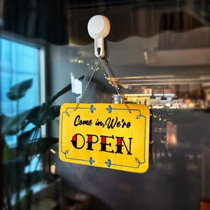 Peut inclure: Une enseigne rectangulaire jaune avec les mots "Come in, We're OPEN" en lettres noires et rouges. L'enseigne est suspendue par une chaîne à une ventouse blanche. L'enseigne a une bordure bleue et des éléments décoratifs.