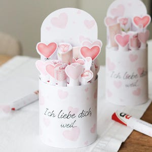 以下が含まれることがあります： ピンクのハートと「Ich liebe dich weil...」の文字で装飾された2つの白い円筒形の容器には、ピンクのハートとギフト型の切り抜きが詰められています。容器はアーチ型のトップで、白い表面に置かれています。
