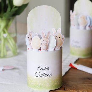 Puede incluir: Decoración de Pascua cilíndrica blanca con un degradado verde y amarillo en la base. La decoración presenta imágenes recortadas de conejos, huevos y un pollito. Las palabras "Frohe Ostern" están impresas en la parte delantera.