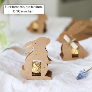 May include: Cardboard bunny-shaped gift boxes with a cutout to hold a chocolate candy. The boxes are brown and have small dot details. The text "F&uuml;r Momente, die bleiben. DIYCarinchen." is in a white box in the upper left corner.