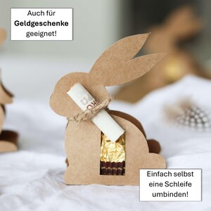 May include: A brown cardboard bunny-shaped gift box with a cutout for treats. A rolled-up paper is tied to the bunny's neck with twine. The box contains a gold-wrapped chocolate. Text on the image says, "Also suitable for money gifts!" and "Simply tie a bow yourself!"
