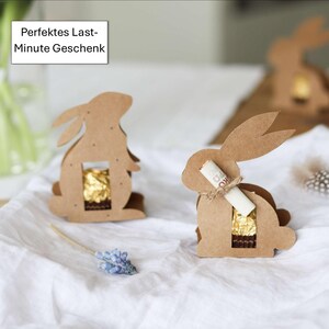 May include: Cardboard Easter bunny-shaped gift boxes. Each bunny has a rectangular opening revealing a gold-wrapped chocolate treat. One bunny has a rolled-up paper scroll tied with twine. The text "Perfektes Last-Minute Geschenk" is in a white box in the upper left corner.
