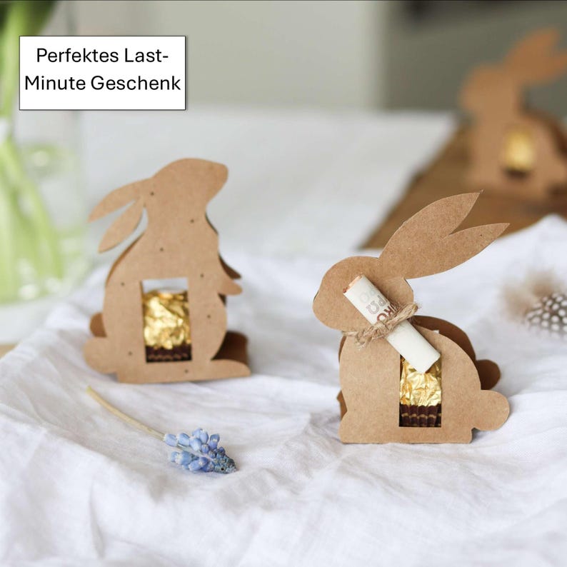 May include: Cardboard Easter bunny-shaped gift boxes. Each bunny has a rectangular opening revealing a gold-wrapped chocolate treat. One bunny has a rolled-up paper scroll tied with twine. The text "Perfektes Last-Minute Geschenk" is in a white box in the upper left corner.