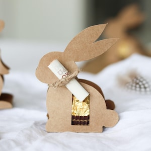 以下が含まれることがあります： 金色のチョコレートが見える切り抜き部分がある茶色の段ボール製のイースターバニー型のギフトボックス。丸められた紙の巻物が紐でウサギに結び付けられています。