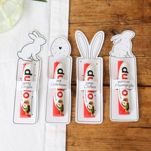 Könnte beinhalten: Vier Oster-Geschenkanhänger mit einer rot-weißen Süßwarenriegel. Jeder Anhänger hat ein anderes Design: ein Hase, ein Ei und Hasenohren. Die Anhänger tragen deutschen Text: "Hasige Grüße", "Vom Osterhasen", "Frohe Ostern" und "Hoppelige Hasengrüße".